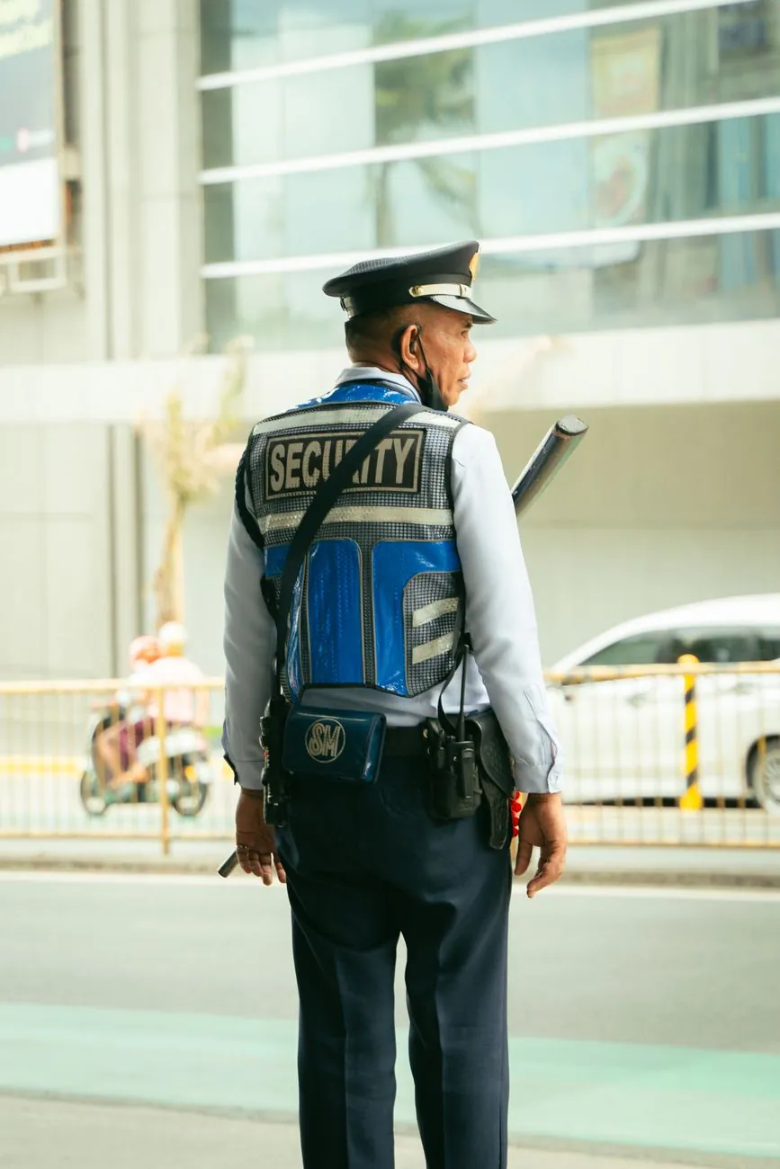 Guardia de seguridad con chaleco y gorra, de espaldas, vigilando la entrada de un edificio