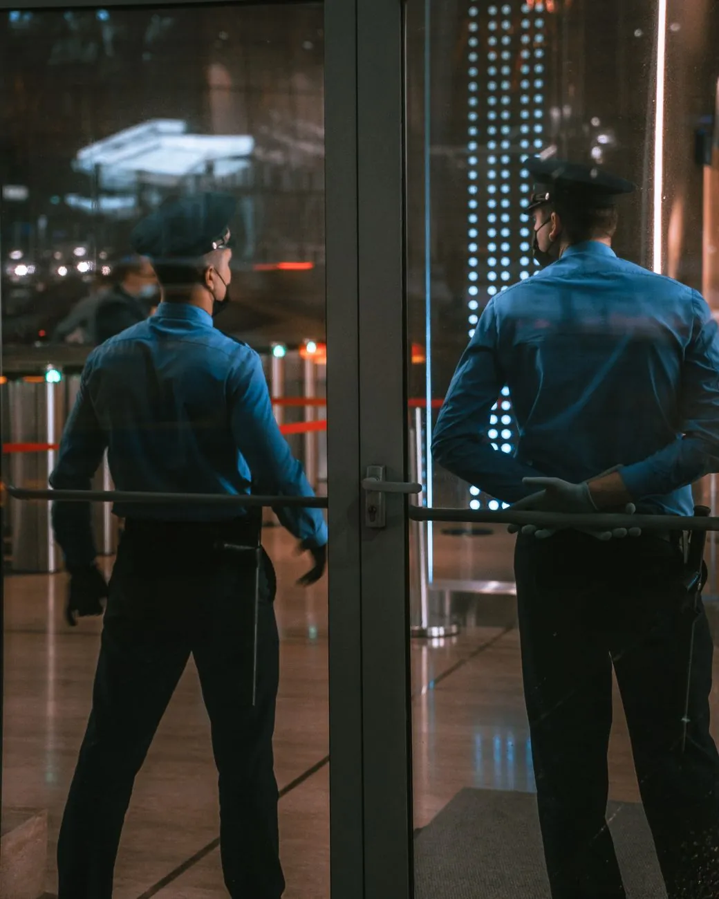 Dos guardias de seguridad de uniforme azul de espaldas frente a la puerta de vidrio de un edificio