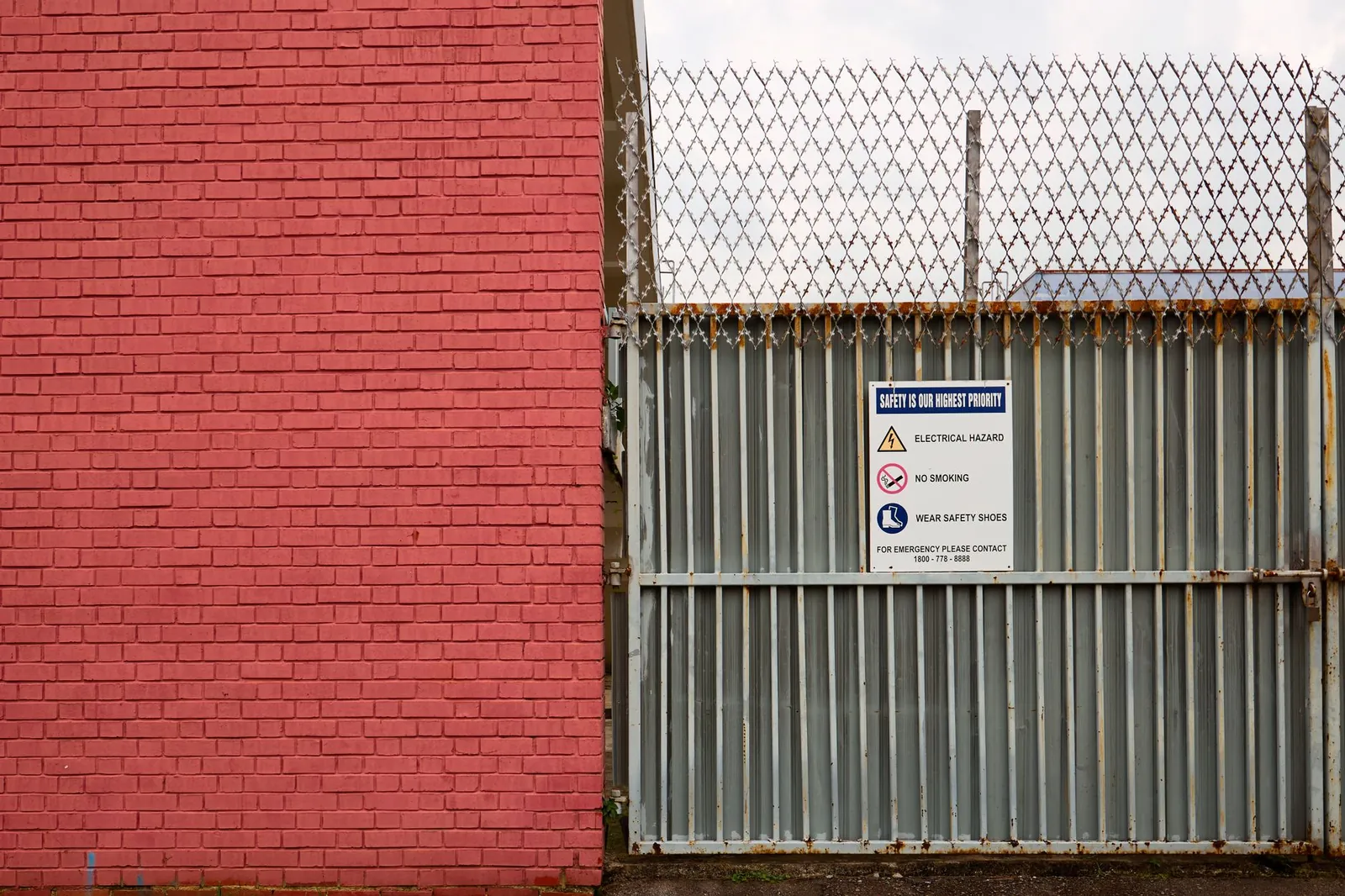 Portón metálico de una instalación industrial con letreros de advertencia junto a un muro de ladrillo